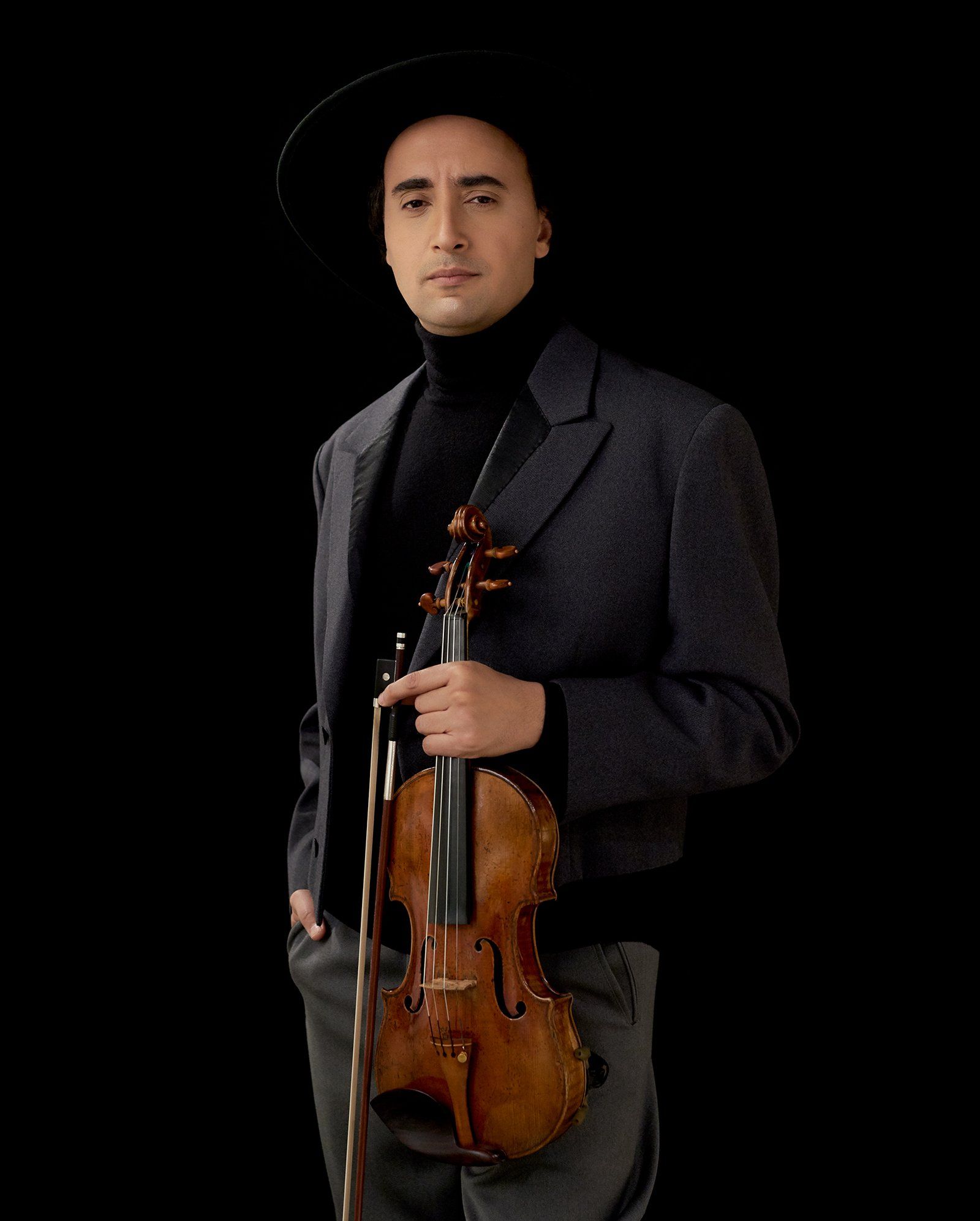 Mario Hossen wearing a wide-brimmed black hat and grey blazer over a black turtleneck, holding his violin and bow, one hand in his pocket