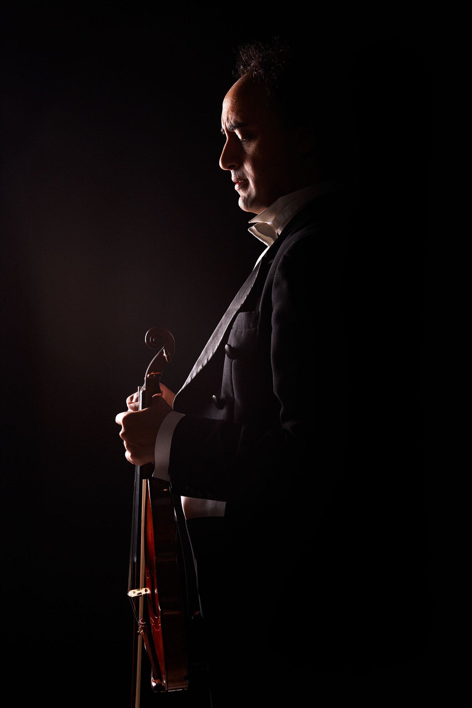 Side-profile silhouette of Mario Hossen in formal black tie, holding his violin and bow vertically, lit with dramatic rim lighting against near-black