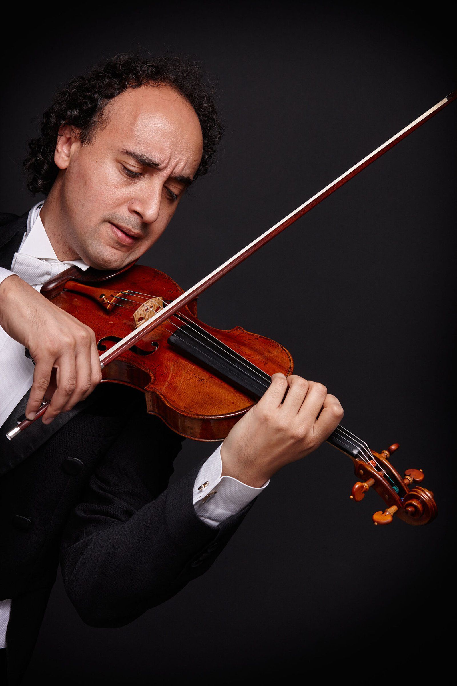 Mario Hossen playing the violin in profile, white bow tie and shirt cuff visible beneath a dark formal jacket, the instrument lit against black