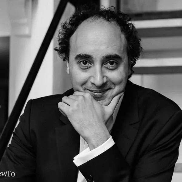 Black-and-white candid portrait of Mario Hossen smiling, chin resting on his hand, seated indoors near a staircase