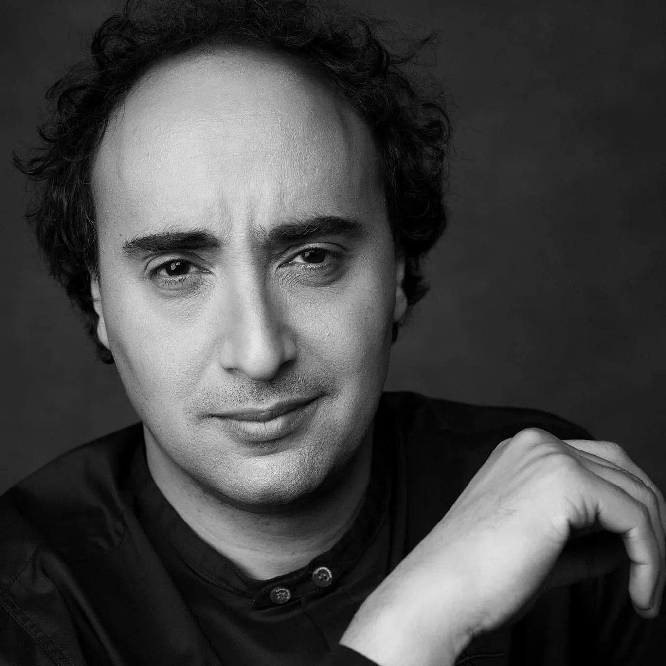 Black-and-white close head-and-shoulders portrait of Mario Hossen, wrist resting near his jaw, looking directly at camera