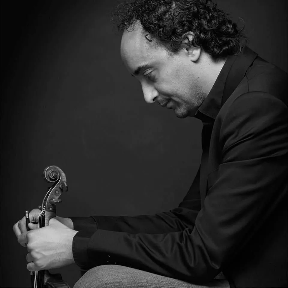 Black-and-white photograph of Mario Hossen seated in profile, adjusting the tuning pegs of his violin, looking down in concentration