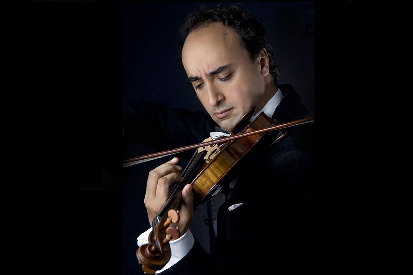 Mario Hossen playing the violin, head bowed over the instrument, dramatic side-lit studio portrait against a black background