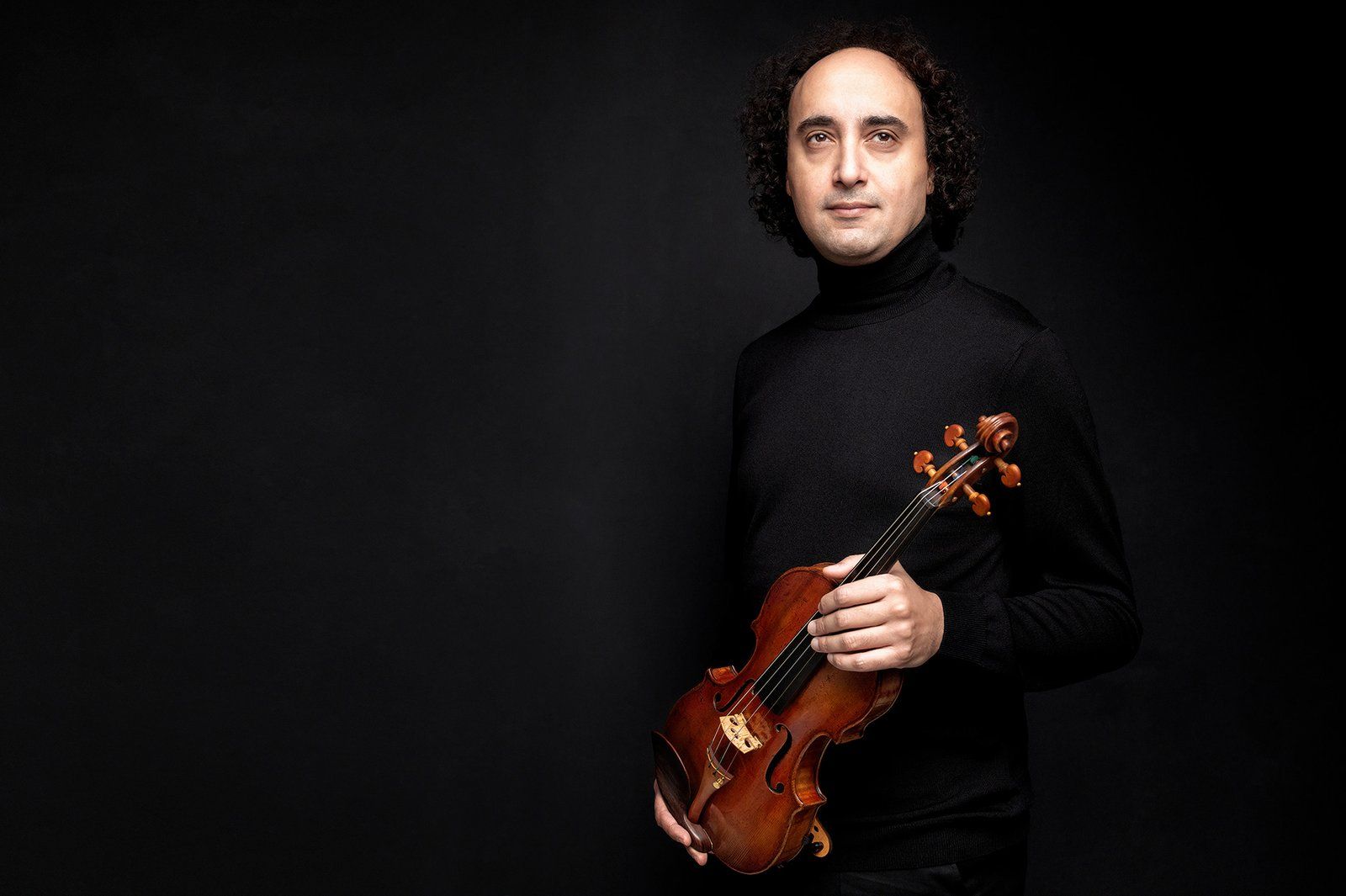 Mario Hossen in a black turtleneck, standing three-quarter view, holding his violin by the neck at chest height, looking directly at camera
