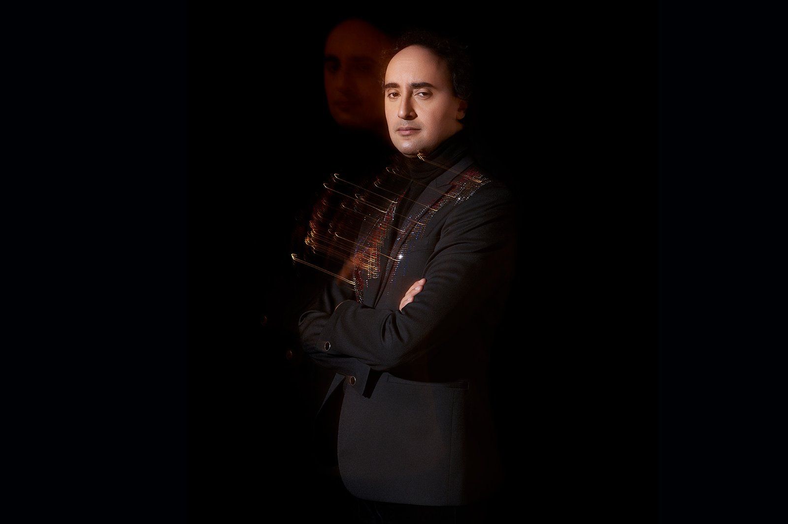 Mario Hossen with arms crossed in a dark embellished jacket over a black turtleneck, a faint ghosted profile visible in the shadow behind him