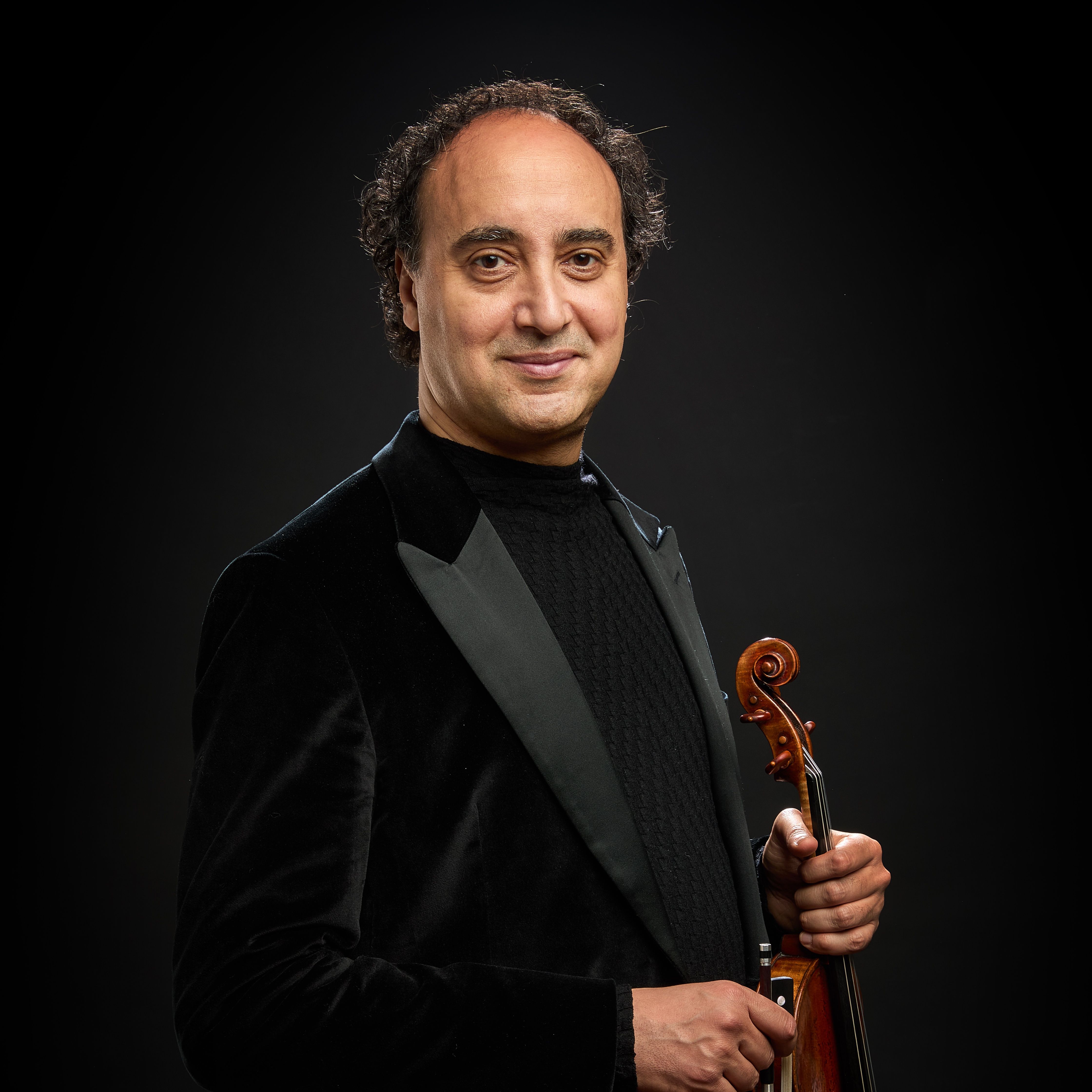Mario Hossen in a black velvet tuxedo jacket, holding his violin upright by the neck with the bow, three-quarter studio portrait against a black background